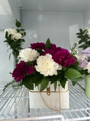 Magenta and white carnations with ruscus in a white quilted handbag-shaped container with a gold chain strap, sitting on a cooler shelf.