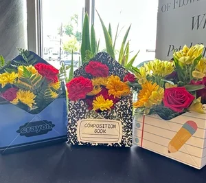 Three teacher-appreciation pieces on the shop counter by the window: flowers in a composition-book box, a crayon box and a notebook-paper box with a pencil graphic.