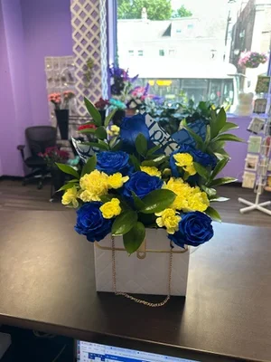 Blue-dyed roses and yellow carnations in a white quilted handbag-style container with a gold chain handle, mylar balloon tucked in, sitting on the shop counter.