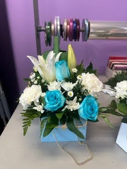 Turquoise-dyed roses with white lilies, carnations and daisies in a light blue novelty purse box with a gold chain, on the shop workbench under the ribbon spools.