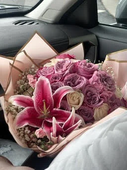 Hand-tied bouquet of mauve roses and stargazer lilies in blush paper wrap, sitting on a car seat mid-delivery