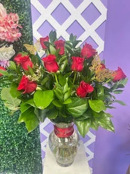 Dozen red roses with greenery in a clear glass vase, shot against the boxwood and white lattice photo backdrop