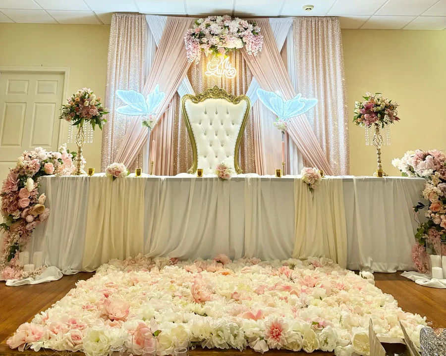 Head table with cream draping, two gold high-back chairs, tall floral columns and a carpet of pink and white blooms across the floor.
