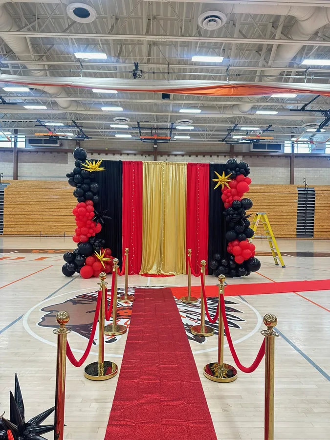 School gymnasium entrance with a red carpet, gold stanchions and red, white and blue balloon columns.