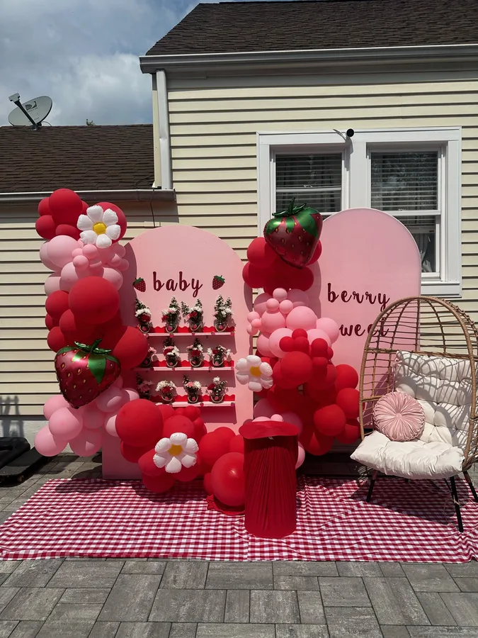 Outdoor baby shower with a strawberry theme: red and pink balloon garland, a hanging chair and a gingham floor.