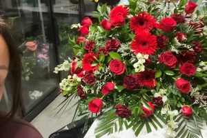 Wide one-sided all-red spray (roses, gerbera, carnations) fanned with palm and leatherleaf, photographed on the shop counter in front of the glass flower cooler.