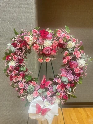Open heart wreath in pink, lavender and hot pink on a black wire easel, finished with a white satin bow and pink butterflies, against a grey funeral-home wall.