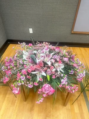 Full casket spray of pink lilies, roses, carnations and stock on a low stand in a funeral-home room with grey grasscloth walls and a wood floor.