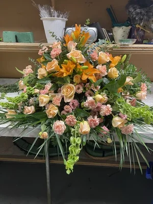 Peach, orange and green casket spray (lilies, roses, carnations, bells of Ireland) on a stand in the workroom, with floral-foam bricks and shop shelving behind.