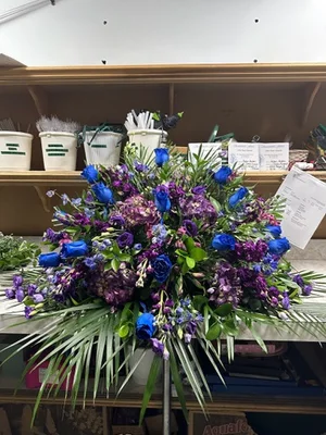 Royal blue and purple casket spray on a stand in the workroom, with vase buckets, order slips and the shop bench behind it.