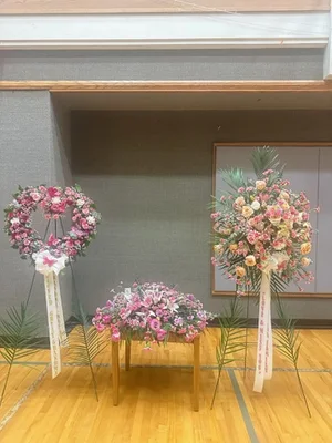 Funeral-home room shot with three pieces together: the pink heart on an easel, the pink casket spray on a stand, and a peach standing spray on an easel with a printed condolence sash.