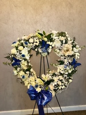 White open heart wreath (roses, lilies, mums, baby's breath) with blue butterflies and a royal blue satin bow, on a black wire easel against a plain tan wall.