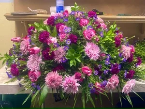 Large pink, magenta and purple casket spray (roses, dahlias, carnations, asters) on the workroom bench, with the shop's shelf and supplies behind it.