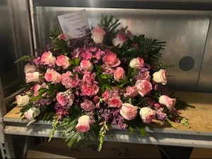 Pink and cream rose casket spray on a metal cooler shelf, with a handwritten funeral order card tucked into the top of the arrangement.