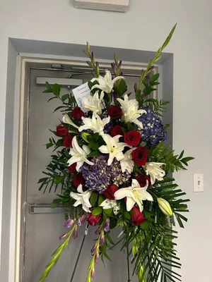 Tall standing spray of red roses, white lilies, blue hydrangea and gladiolus on a black wire easel, card tucked in, shot in front of a metal door.