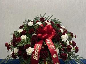 Burgundy and white carnation casket spray with a red satin 'Loving Mother' ribbon sash, against the cooler wall.