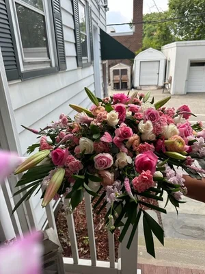 Large pink, coral and white rose-and-lily spray being carried by hand out of a house, photographed against the white siding and porch rail.