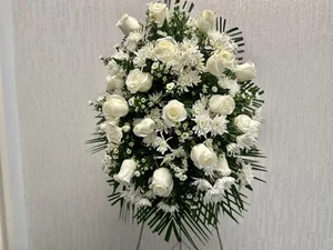 All-white standing spray — white roses, mums and daisies with palm — on a wire easel against a pale textured wall.