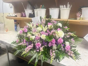 Pink, lavender and white casket spray (stock, snapdragon, carnations, roses, lilies) on the florist's work bench, with design buckets and tools on the shelf behind and an enclosure card tucked in.