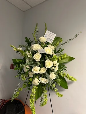 White rose and green standing spray with bells of Ireland trailing, on a wire easel in a room corner, two sympathy cards tucked into the face.