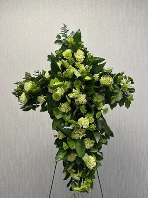 Standing cross of white and green carnations with heavy foliage, on a wire easel against a gray textured wall.