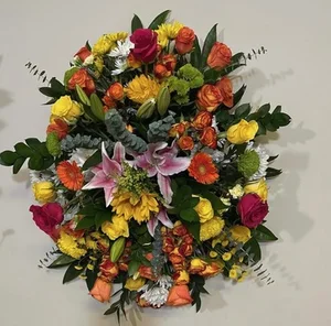 Large all-around oval arrangement in hot orange, yellow and magenta — roses, gerberas, mums, stargazer lilies — sitting directly on a pale studio table with no visible vase.