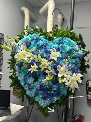 Solid heart of turquoise and blue dyed carnations and delphinium with white lilies, on a wire easel in the shop workroom.