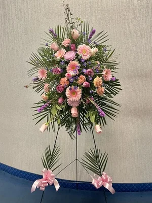 Pink and lavender standing spray — gerberas, carnations, spray roses, statice, palm — on a wire easel with pink bows tied to the easel legs.