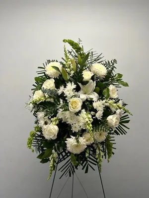White standing spray of roses, mums, lilies and snapdragons with dark greens and palm, on a wire easel against a plain white wall.