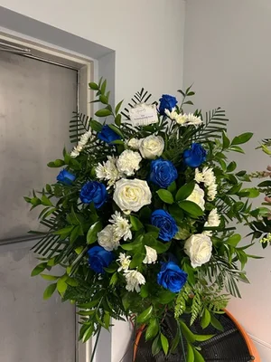 One-sided fan basket of blue-dyed roses and white carnations with an enclosure card, standing in a workroom corner beside a door on a dark orange-trimmed base.