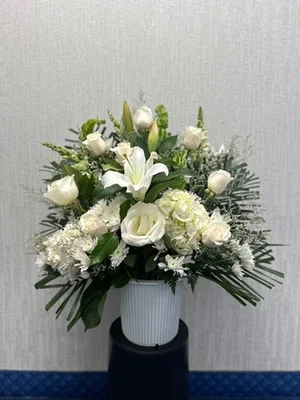 All-white lily, rose, carnation and mum arrangement in a white ribbed funeral urn on a black pedestal, plain wall and blue patterned funeral-home carpet behind.