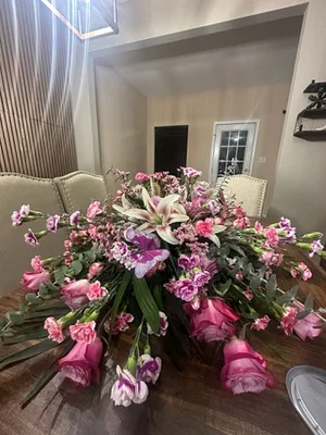 Long low pink piece — lilies, roses, carnations and stock — laid across a dining table with stems trailing over both edges.