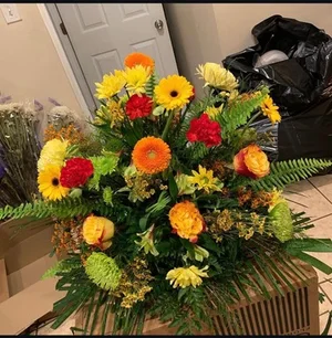 Large one-sided fan basket of yellow and orange gerberas, red carnations, roses and mums on sword fern, shot in the workroom beside flower boxes and trash bags.
