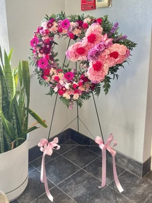 Pink open wreath with a carnation-and-rose butterfly worked across it, on a tall wire easel with pink bows tied to the legs, in a tiled lobby.