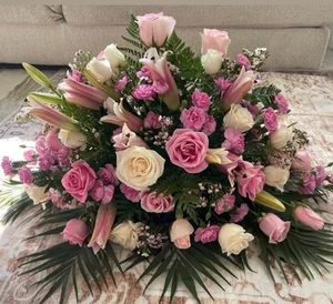 Large pink and cream oval spray of roses, lilies and carnations on a bed of palm fronds and leatherleaf, photographed on a rug in front of a couch.