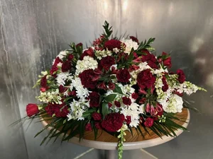 Deep red and white domed oval spray — carnations, roses, snapdragons, hydrangea — on a round wooden table in front of the stainless walk-in cooler.