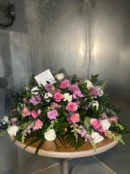 Pink, lavender and white casket spray with an enclosure card on a card pick, on a round table at the walk-in cooler door.