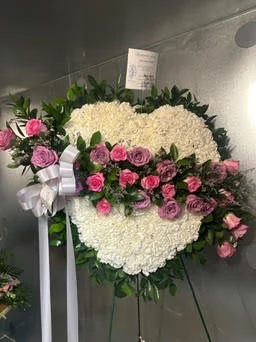 Solid white carnation heart edged in greenery with a pink and lavender rose spray across it and a white satin bow, on a wire easel at the cooler with a sympathy card attached.