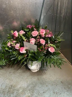Compact pink carnation and rose basket with a white enclosure card tucked in, greenery skirt, sitting on the shop floor by the cooler