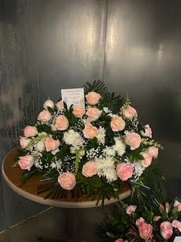 Large pink rose and white chrysanthemum basket with a card, on a round wooden table against the cooler wall