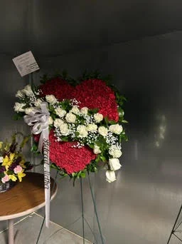 Solid red carnation heart on a wire easel with white rose and carnation accents and a white ribbon, tagged, against the cooler wall