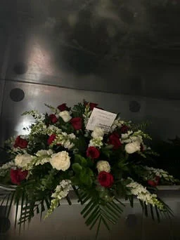 Casket spray of burgundy and white roses with palm fronds and a card laid on top, photographed against the cooler wall