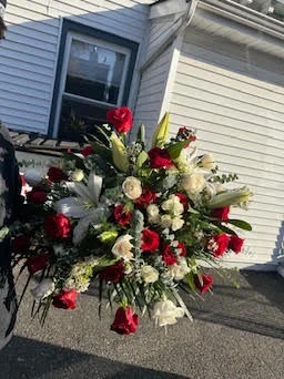 Tall standing spray of red roses and white lilies on an easel, outdoors on a driveway beside a white house, ready for delivery