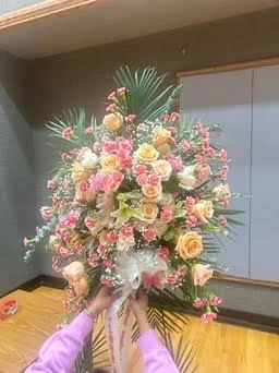 Large fan-shaped spray of peach and cream roses with pink filler and palm fronds, held up by a woman in a lavender sleeve in a wood-floored hall