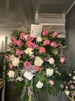 Tall standing spray of pink and lavender roses with white carnations and palm fronds, sympathy cards clipped on, inside the walk-in cooler